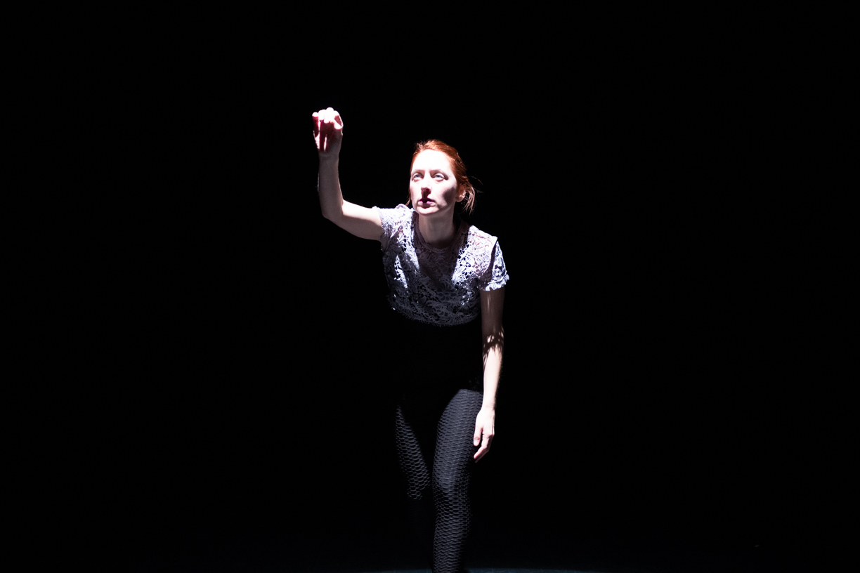 One dancer in a spotlight is writing with her right hand in the air. She has a focused and grave expression on her face.