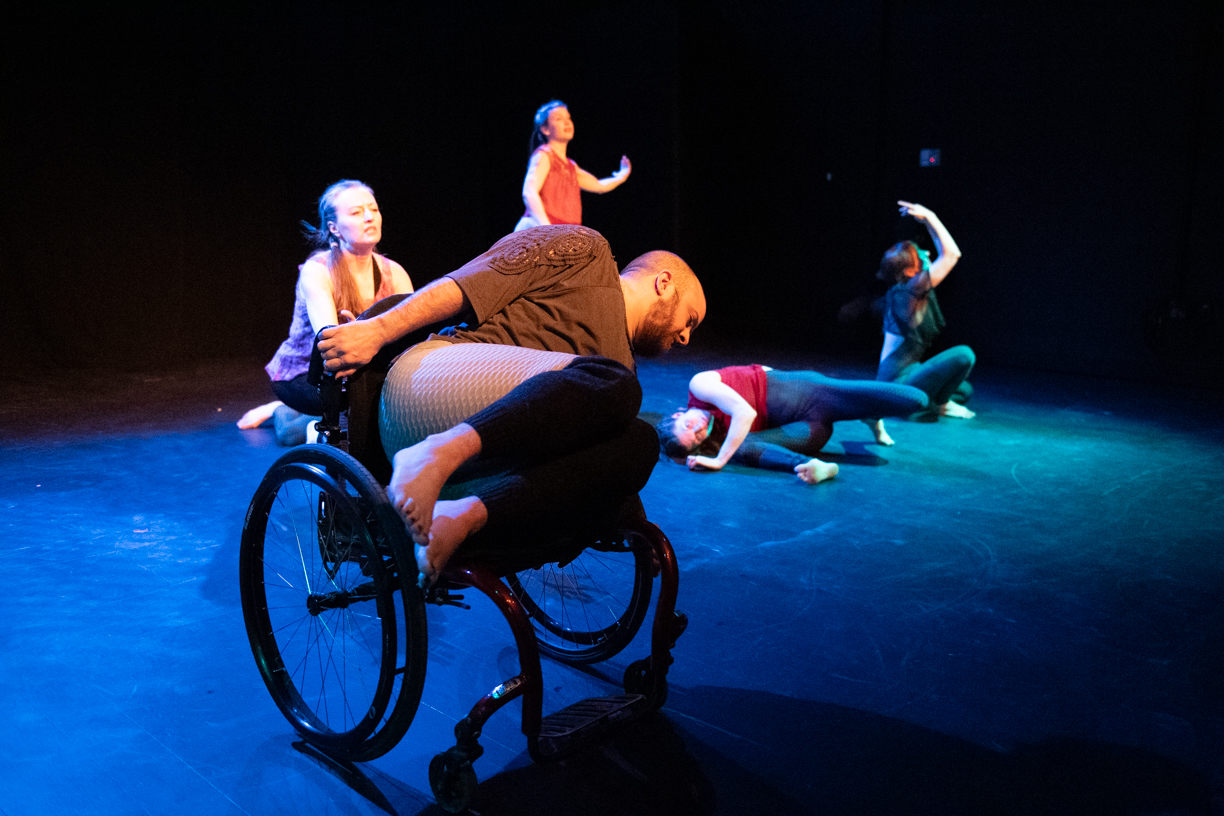 A dancer in a wheelchair is leaning to the left. Thier legs are tucked up and they are lookin gat hte floor. There are four other dancers visible in the background, each doing their own movements.