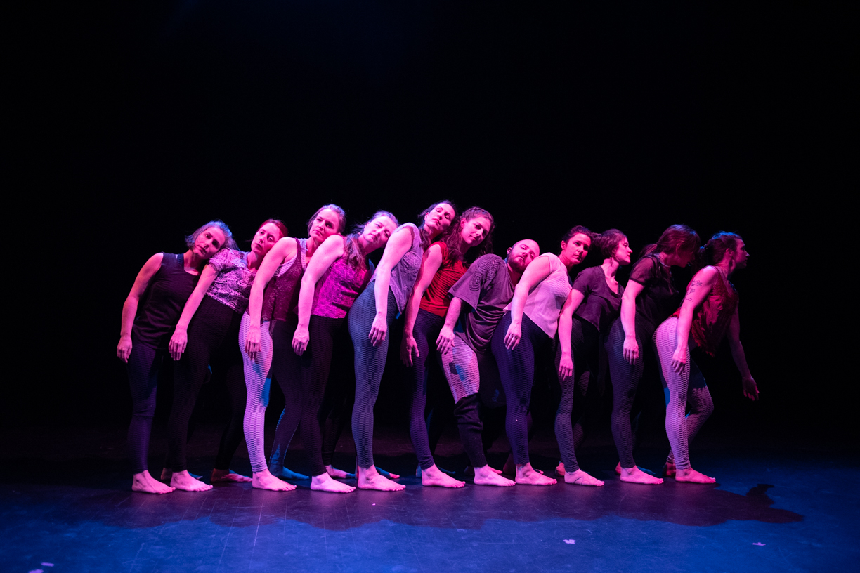 Eleven dancers stand in a close line facing stage left. They are each leaning on the person in front of them, thier heads turned to face the camera.