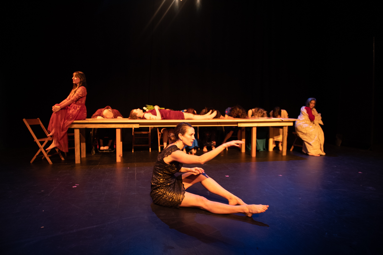 A dancer sits on the floor angled to the left, one leg and one arm outstretched. In the background, a dancer lies on a long table holding flowers. She is bookended by two other dancers sitting at either end. Other dancers in between have their heads down on the table.