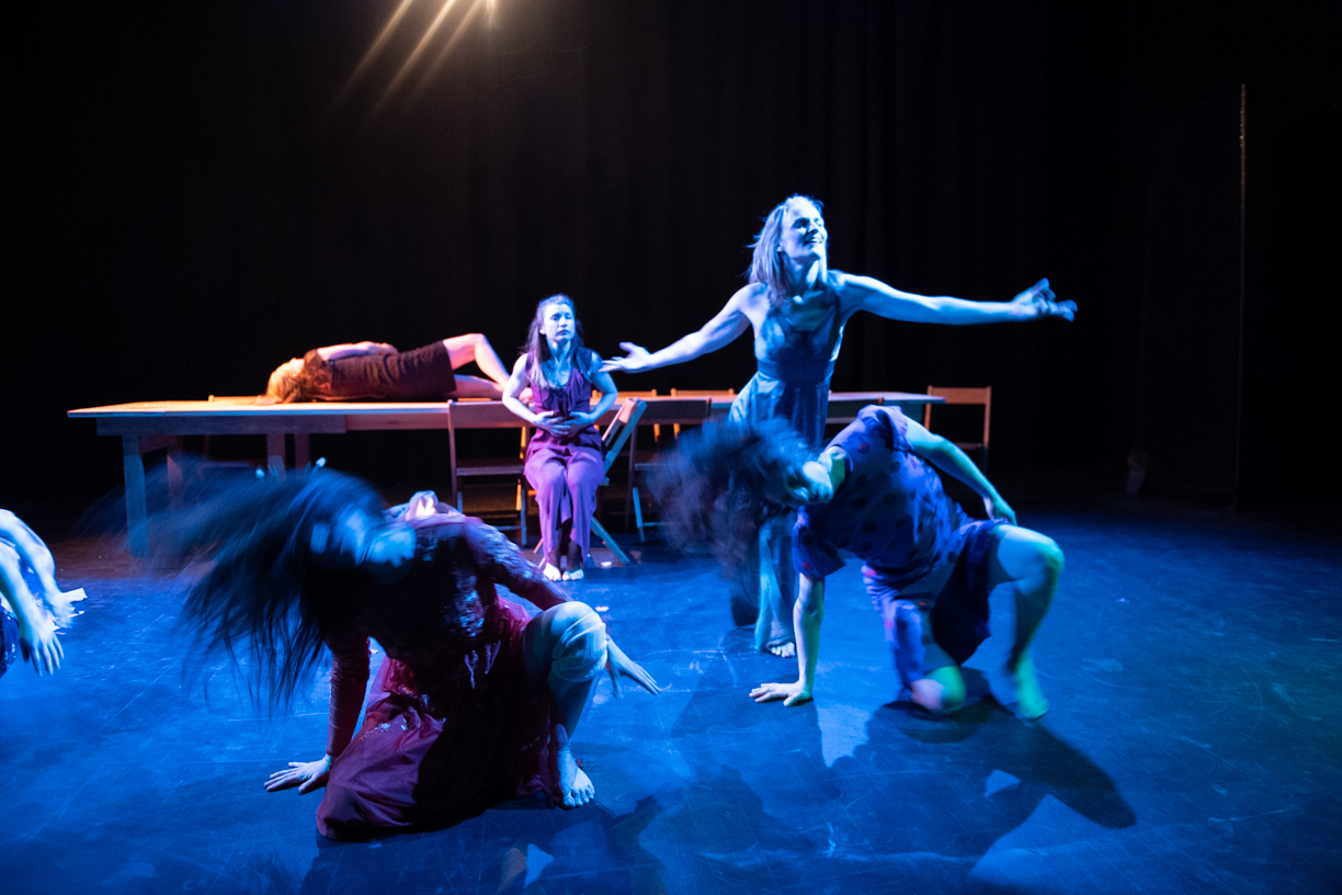 In the foreground, two dancers kneel with thier right hands on the floor. They are swinging their heads to the right and hair is flying. Behind them, a dancers stands with her arms outstretched. Behind her, a dancers sits and looks on. Behind her, a dancer is lying on her back on a long table. 