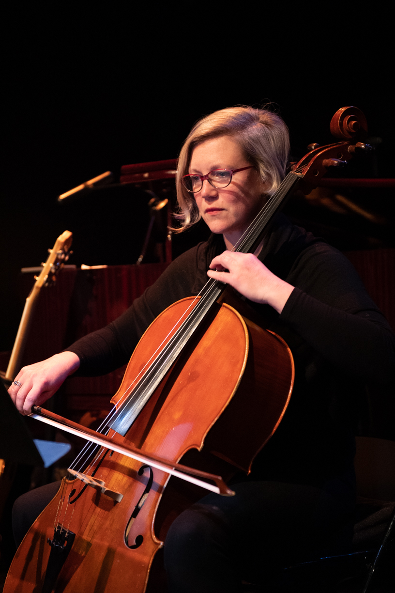 A woman with blond hair and glasses plays the cello. 