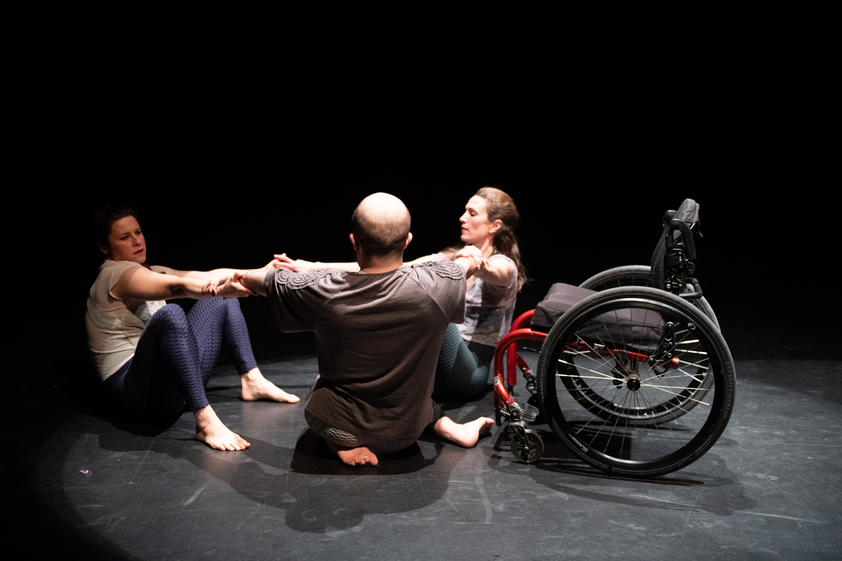 Three dancers sit in a cirlce on the floor holding each others arms. To thier left is an empty wheelchair.