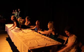 Seven dancers at a dinner table, each lost in their own worlds. In the background, an orchestra can be seen. 
